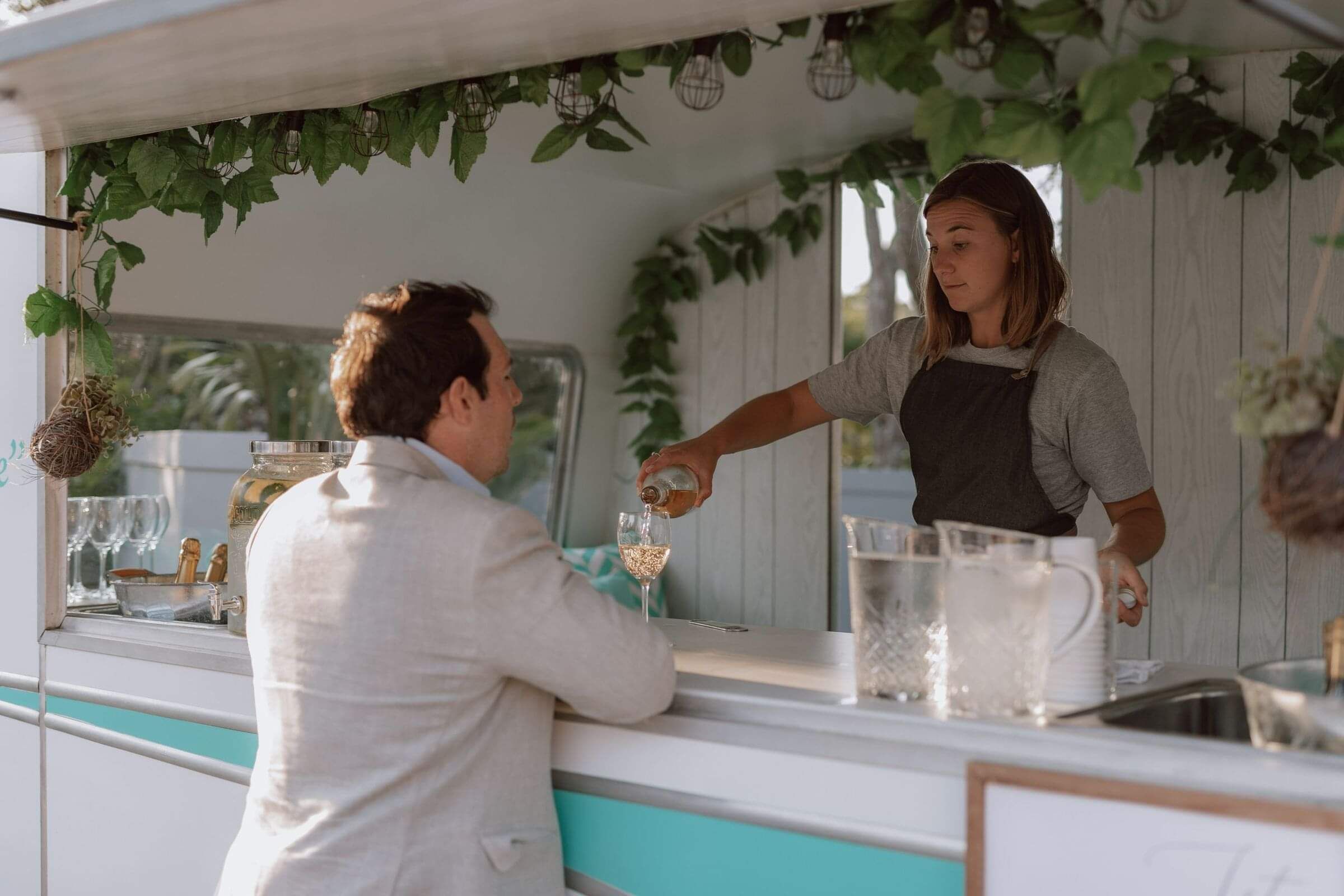 A man standing at the counter of the Vintage Caravan Bar with a girl behind the counter pouring him a glass of wine