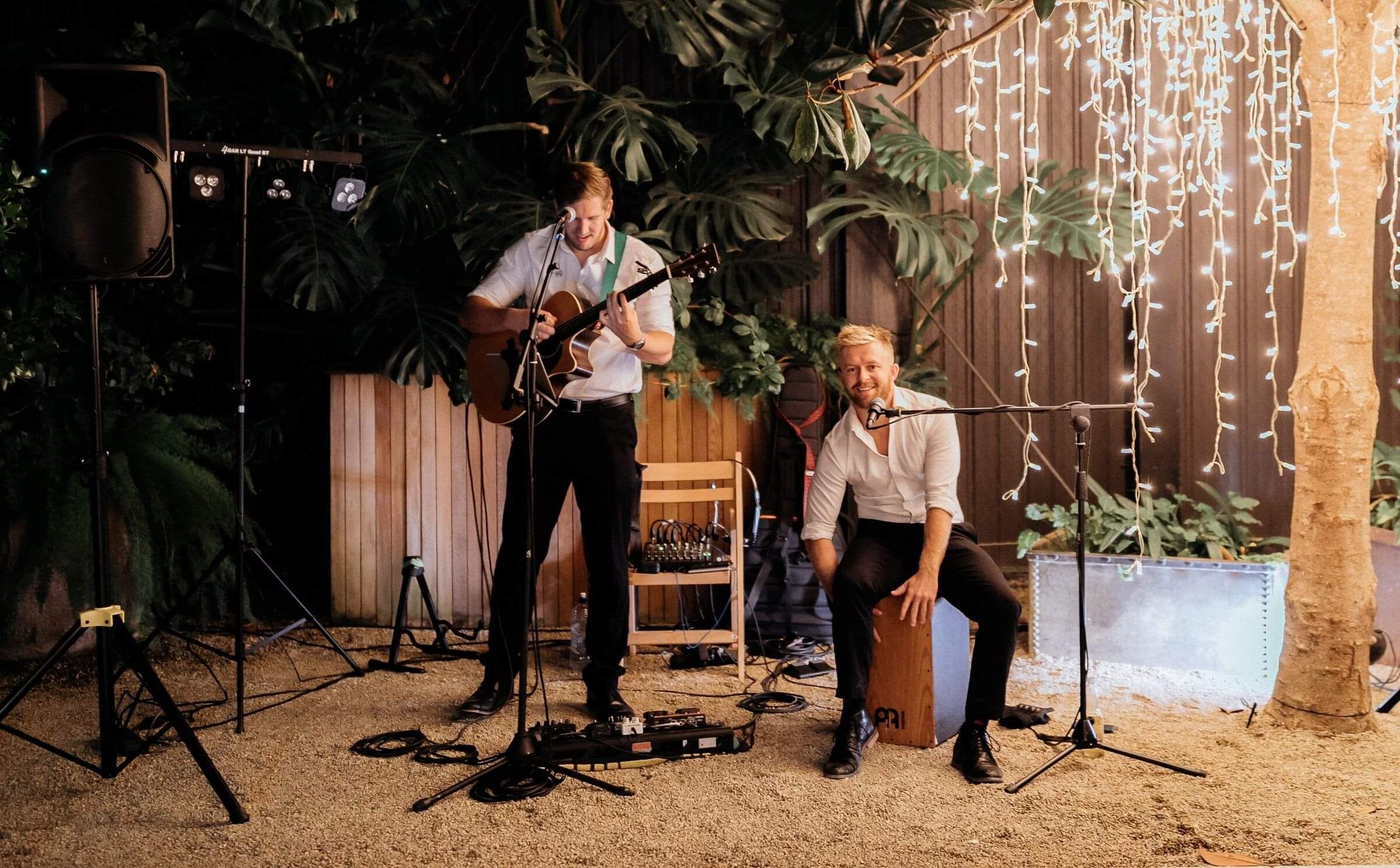 Two men playing in a band, one standing and strumming a guitar and one sitting on a box. Various musical gear behind them.