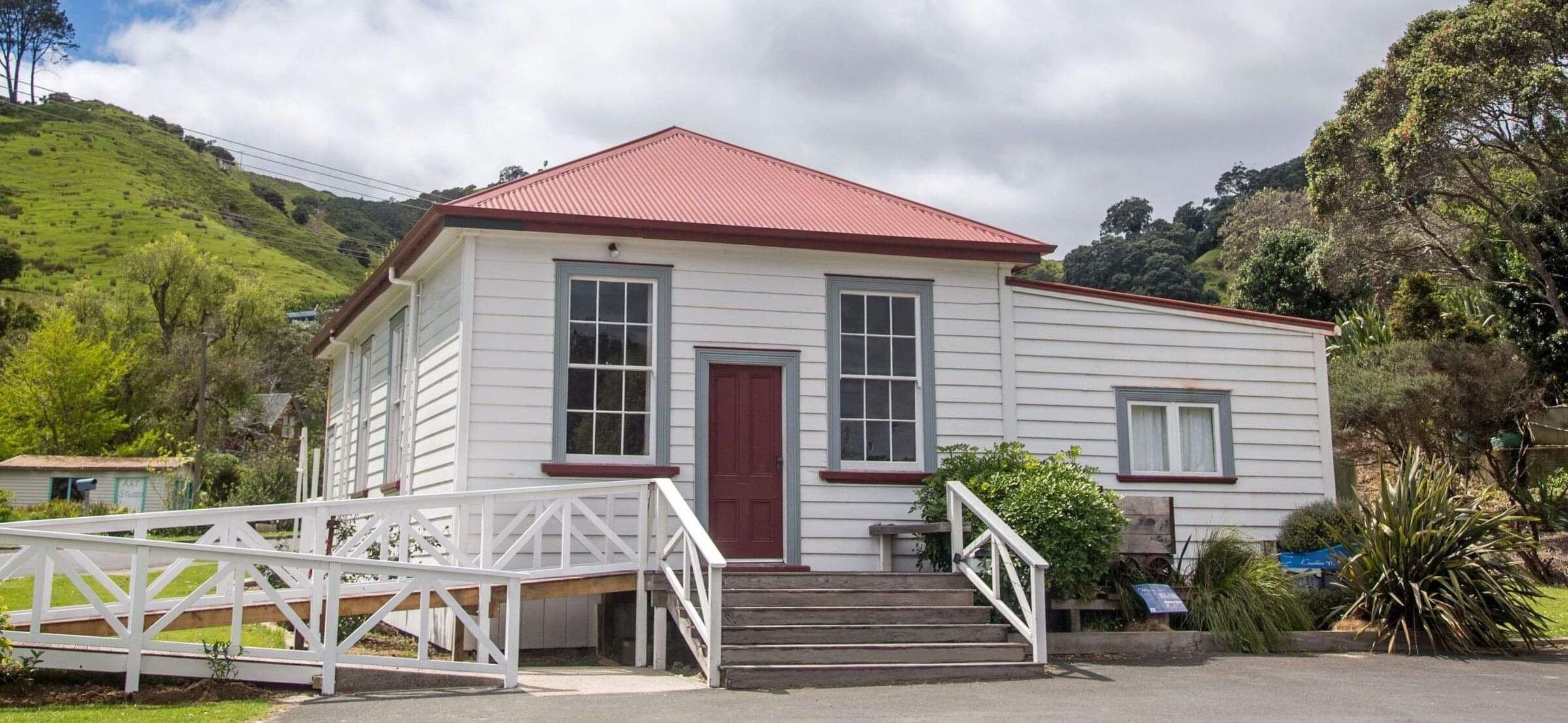 The exterior of the K?aotunu Hall - a white wooden building with a red roof and door, tall sash windows, wooden steps leading up to it and a ramp with