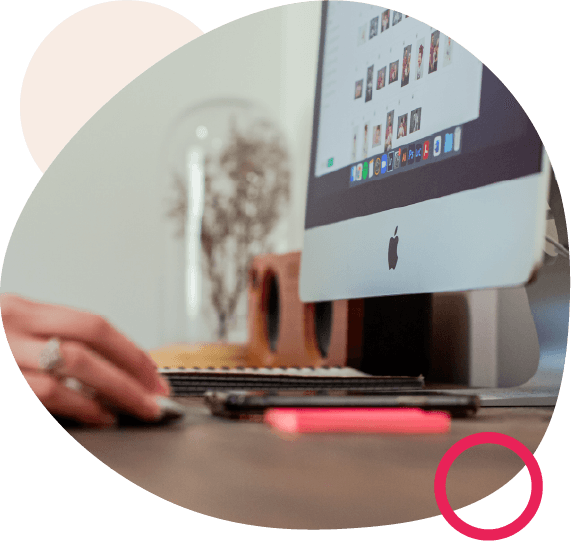 Close-up of a hand using a keyboard at a desk with an iMac, framed by a rounded shape with pink and beige accents