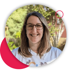Portrait of a woman with glasses and long brown hair, wearing a white t-shirt, standing outdoors, framed by a pink circle