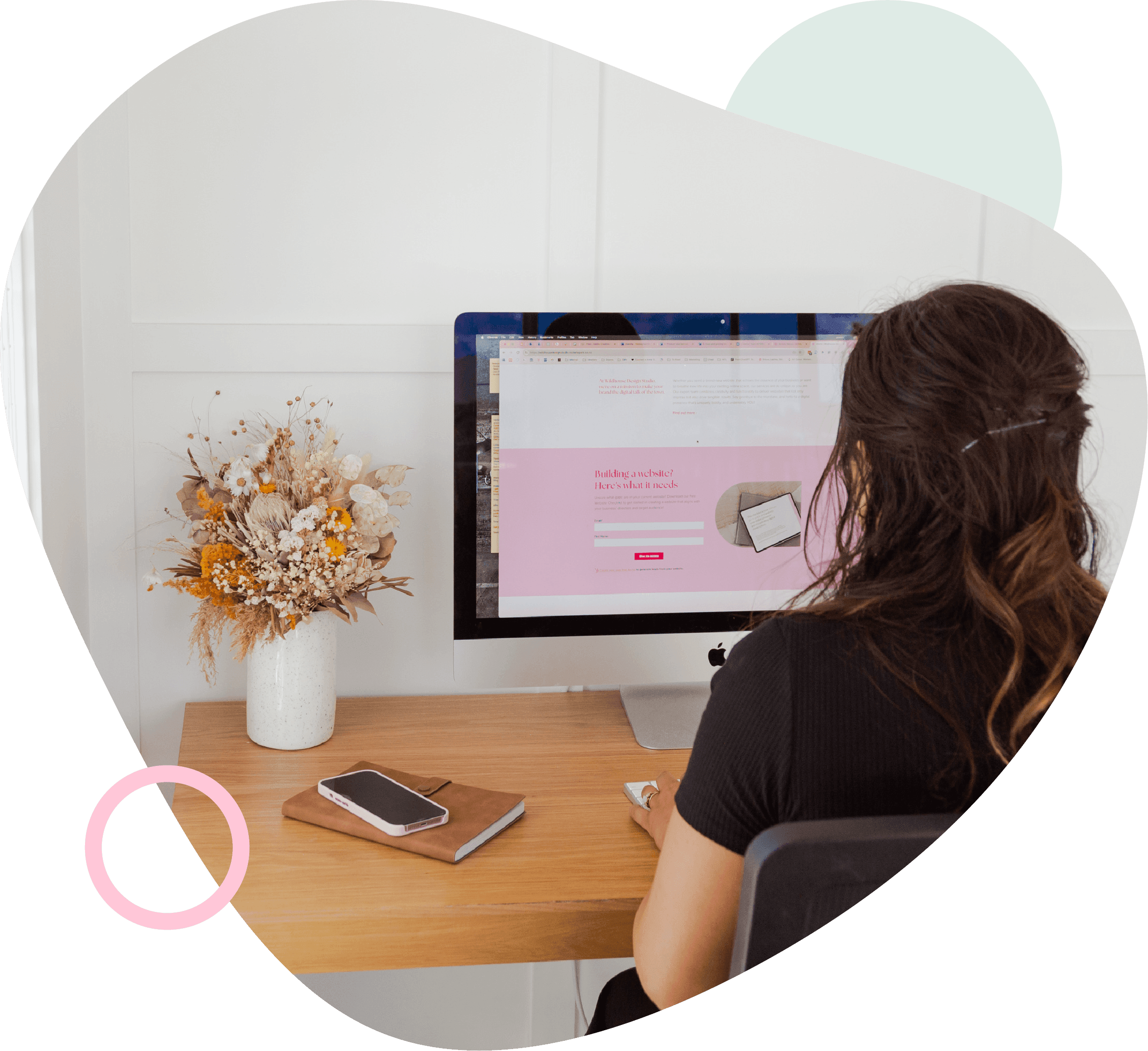 Woman with dark hair working at a desk on a computer, with a phone and dried flowers, framed by a rounded shape with pink and green accents