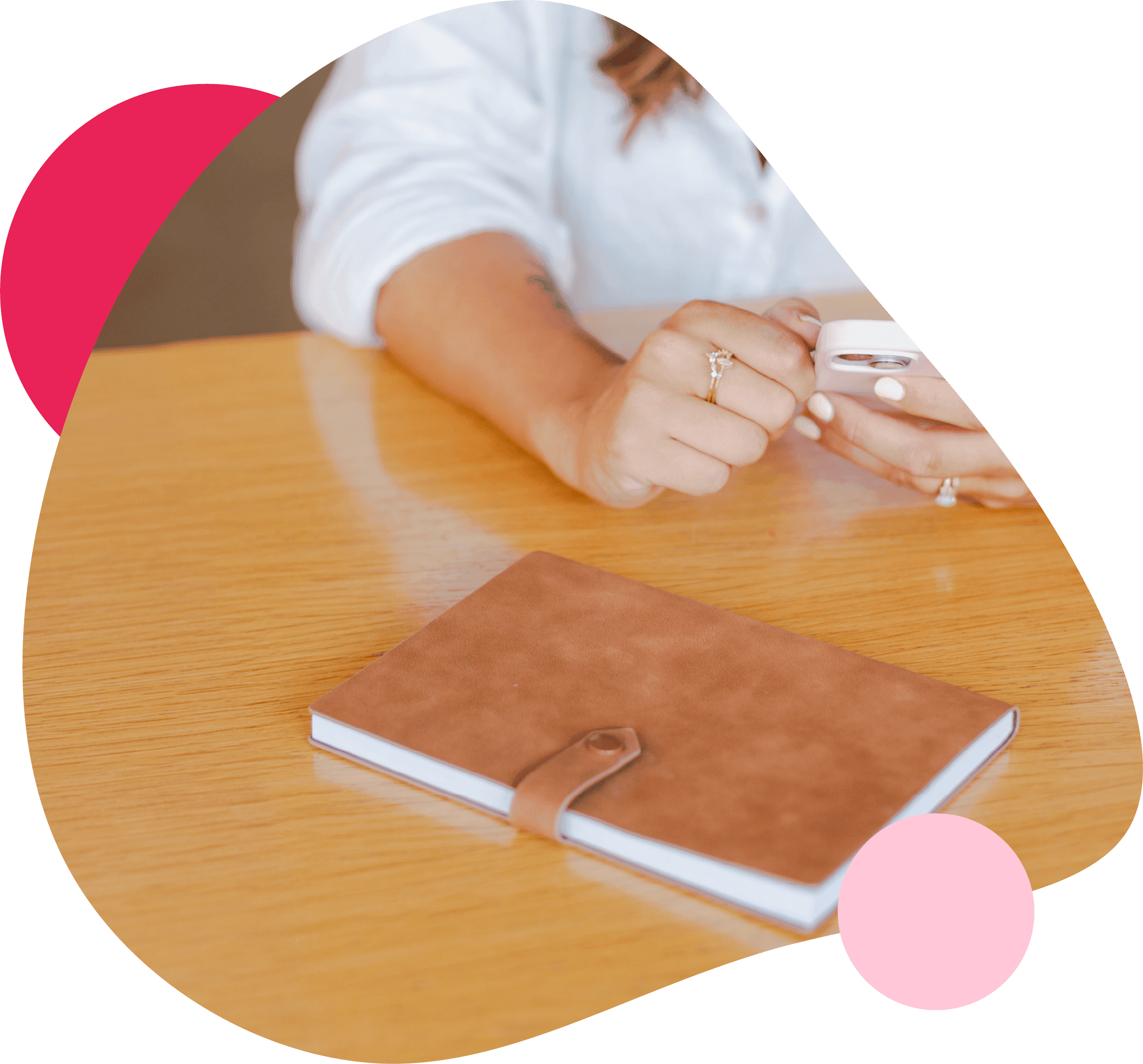 rown leather notebook on a wooden table, with a woman in a white shirt using a smartphone, framed by a rounded shape with pink accents