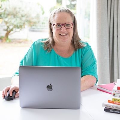 Catherine sitting at a desk using a laptop and mouse, smiling at the camera in a sunny home office.