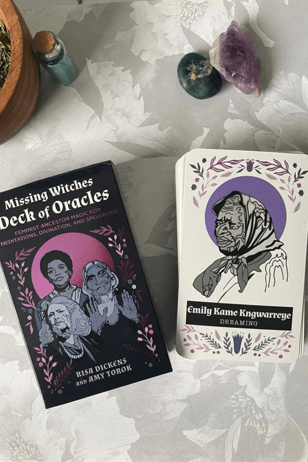 Missing Witches Deck of Oracles pictured the deck and Emily Kame Kngwarrege - Dreaming card on a floral background with cyrstals, glitter bottle