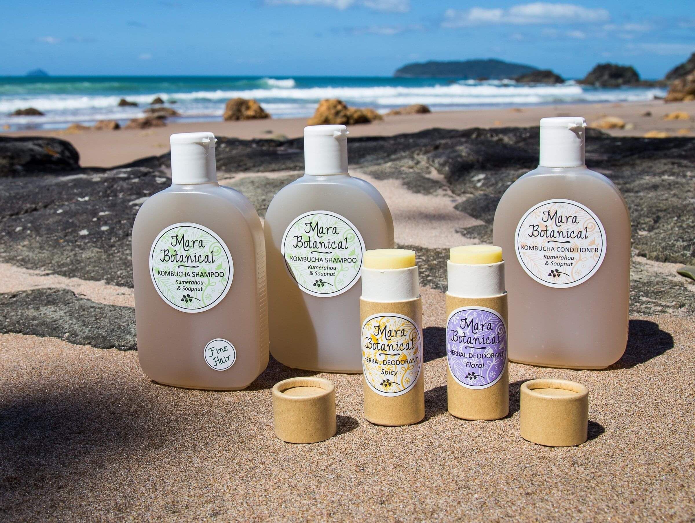 Bottles of handmade hair and body products from Mara Botanical (Kuaotunu) displayed on the sand with the sea behind