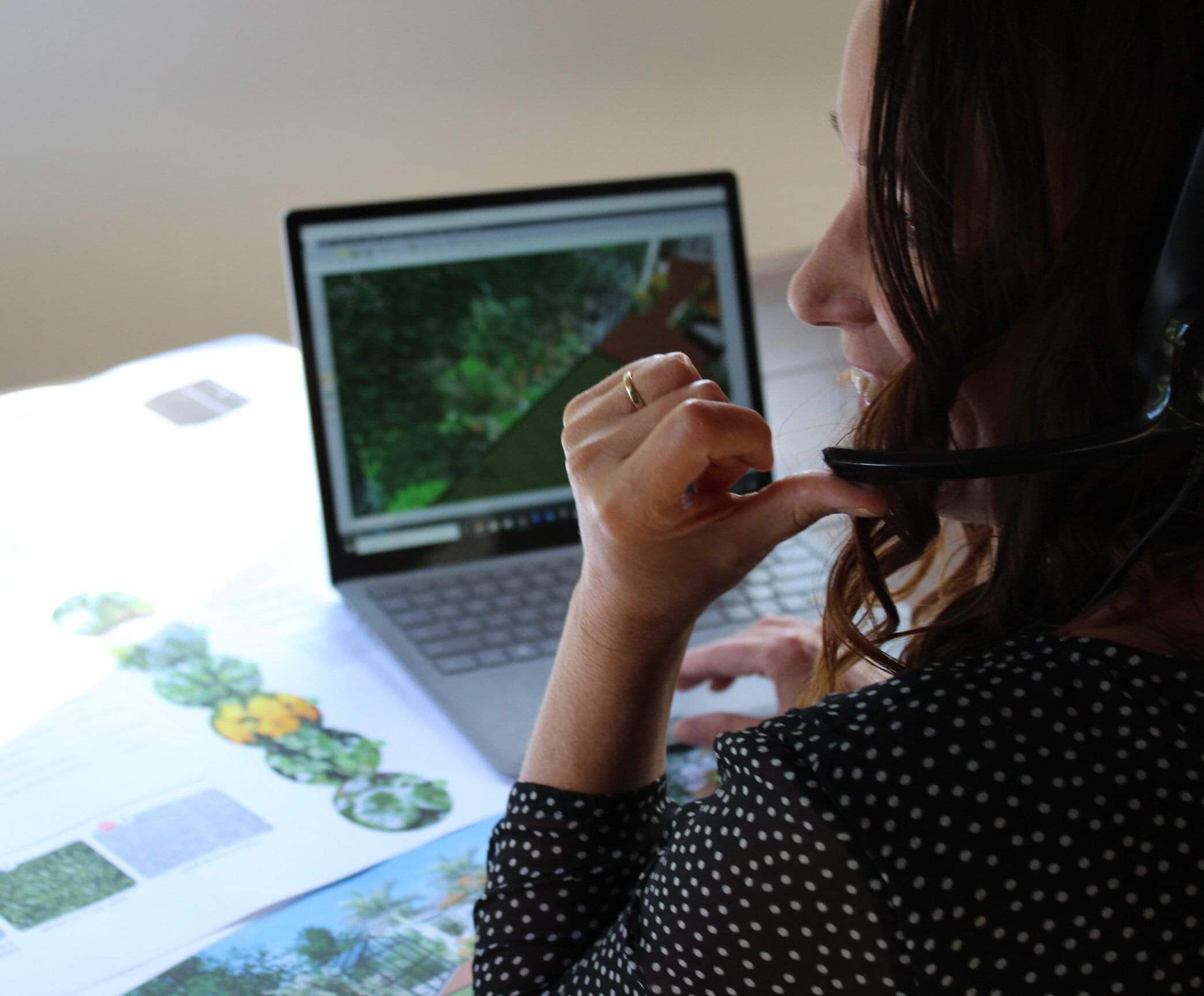 A female landscape designer wearing a headset. She has long, wavy brown hair and sits behind a laptop. Shes looking side on and touching the microphon