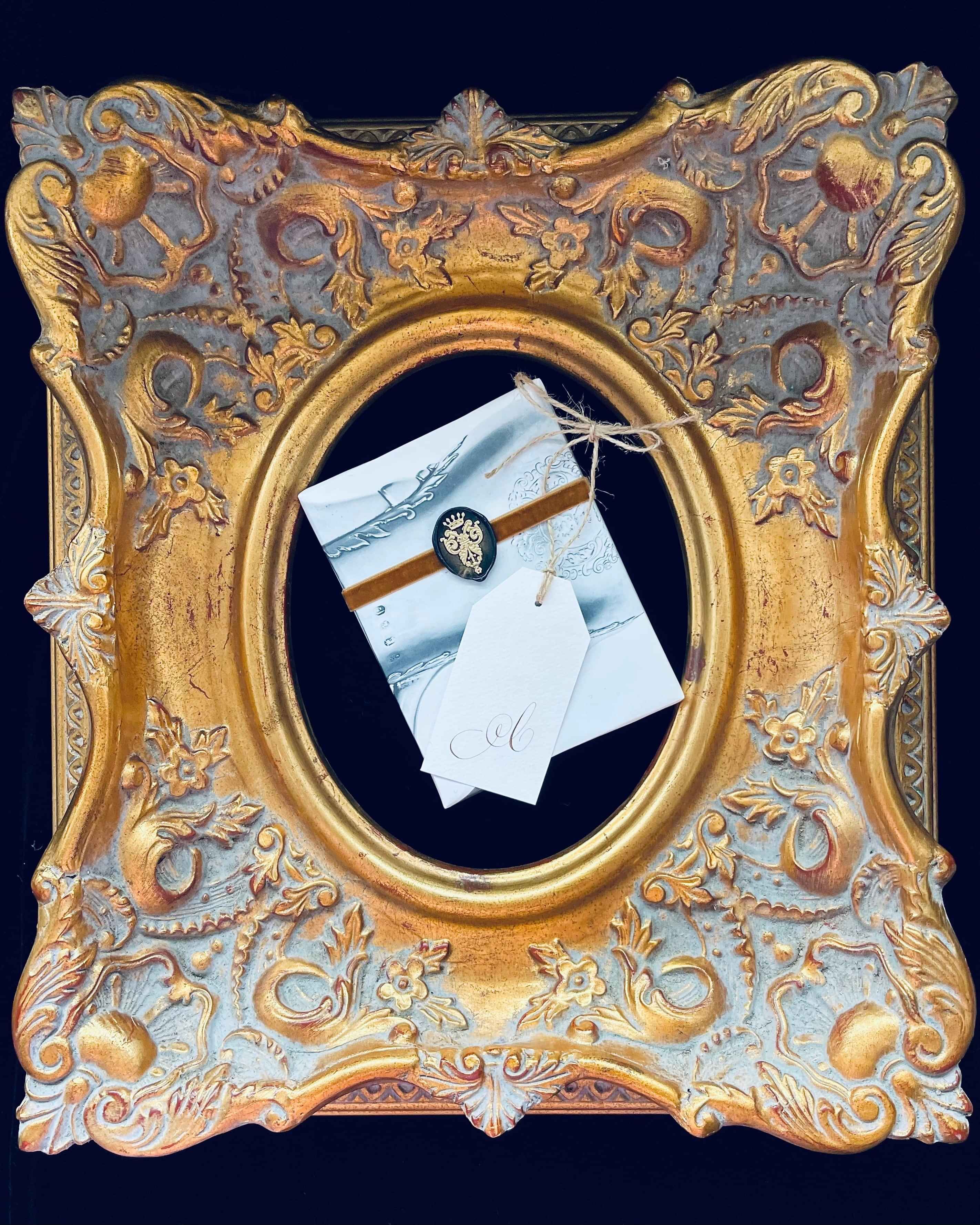 Gilded frame holding a gift wrapped in music sheet paper, gold velvet ribbon, wax seal, and a gift tag with gold copperplate 'C'.