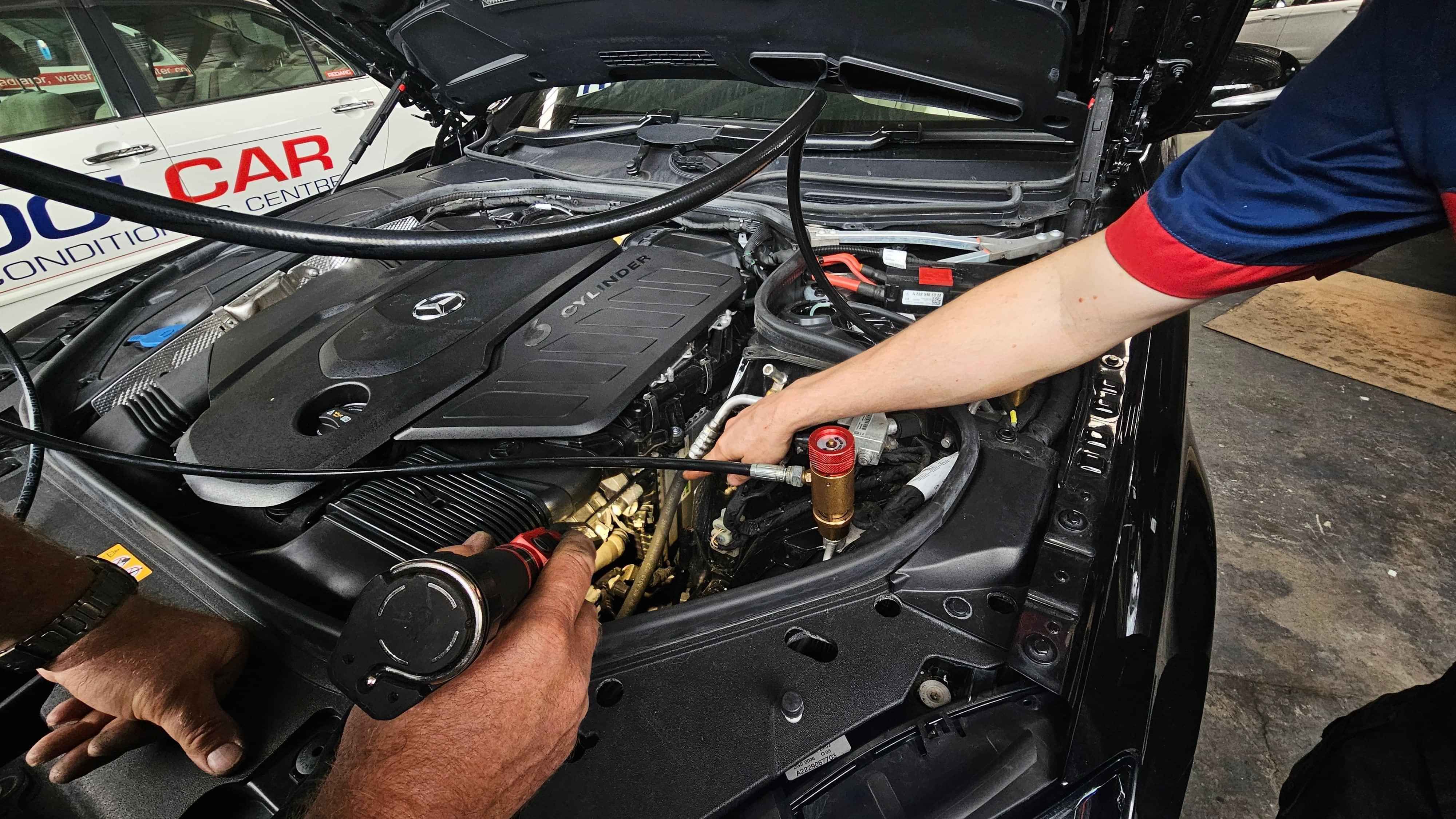 Two technicians at CoolCar Hamilton examining engine bay of S-Class Mercedes for R744 refrigerant leaks using torch.
