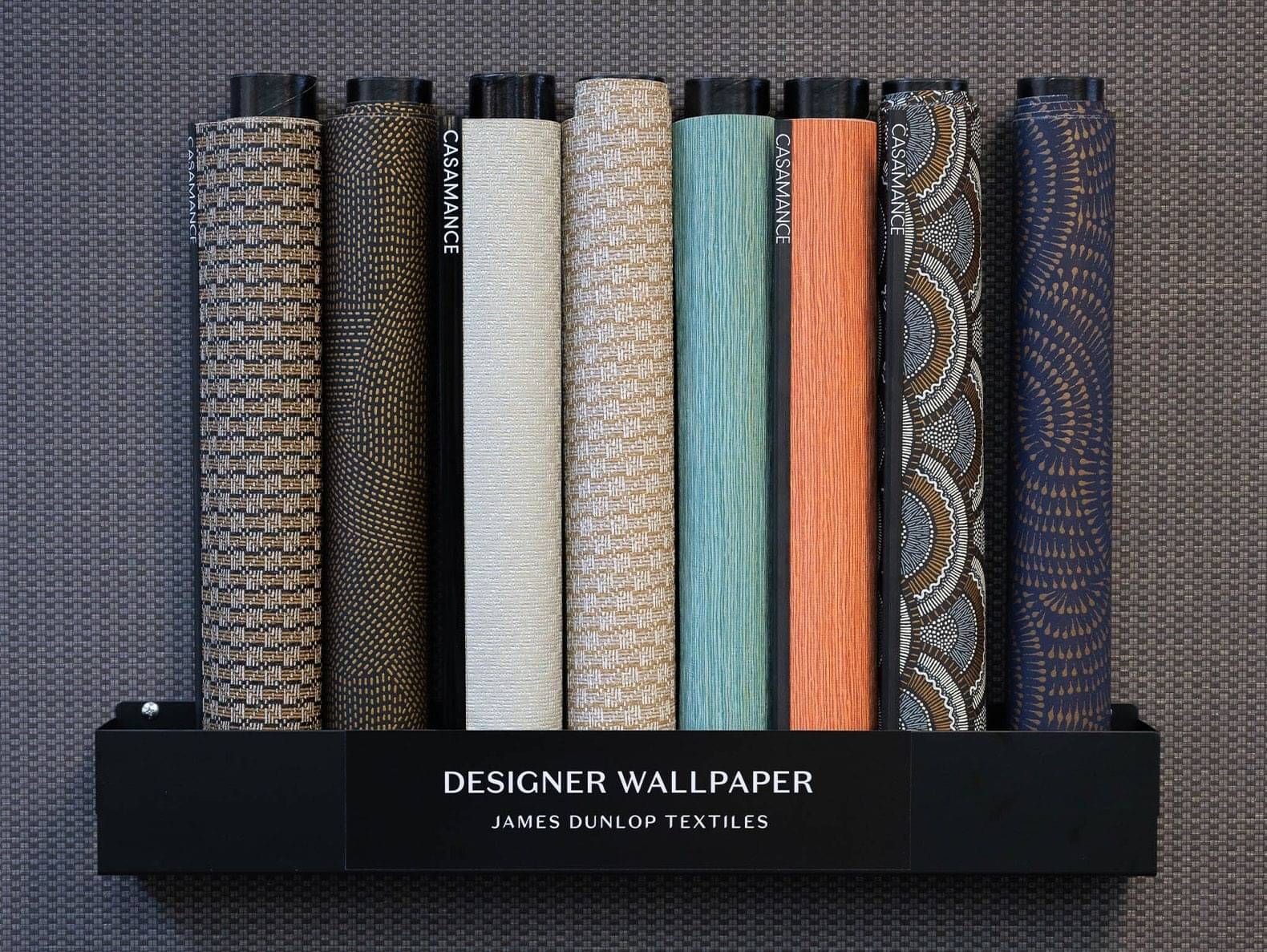 Display of designer wallpaper rolls in various patterns and colours, including gold, white, teal, and orange, from James Dunlop Textiles