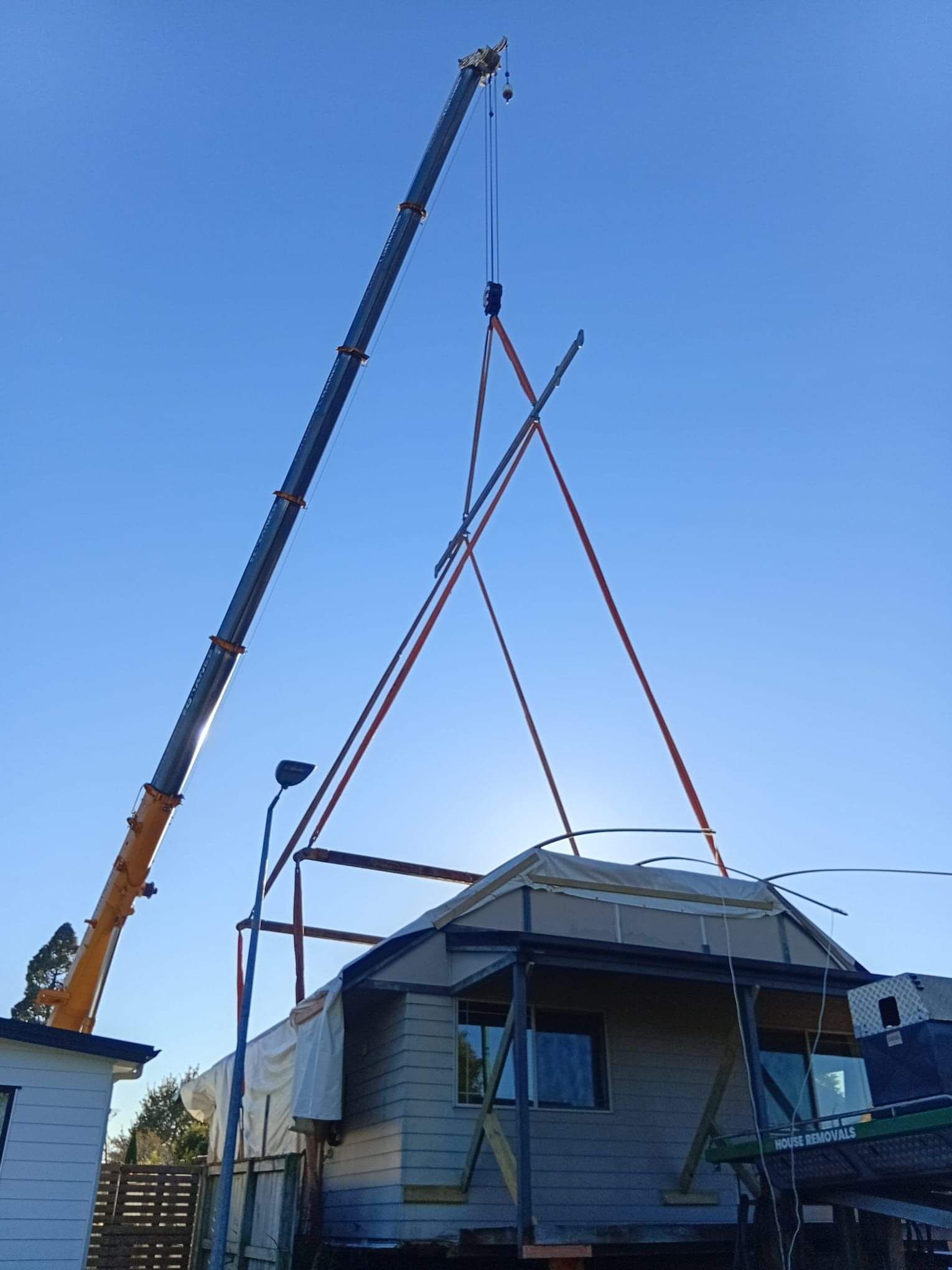 Weatherboard villa suspended on crane spreader bars above a driveway, WBR crew guiding it with a tag line