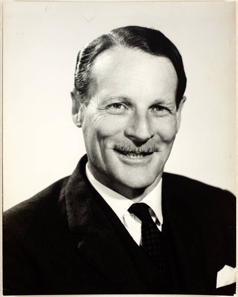 Formal photo in black and white of a man in suit and tie. White pocket square. He has a moustache and is smiling at the camera.