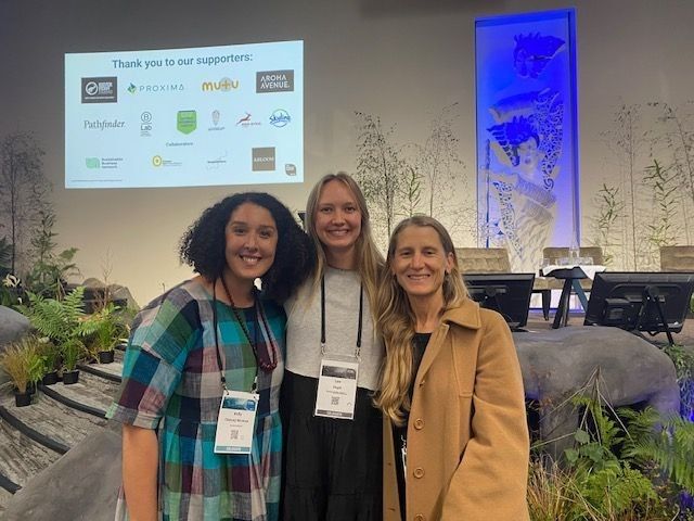 Three attendees smiling at a sustainability or business event in front of a presentation display
