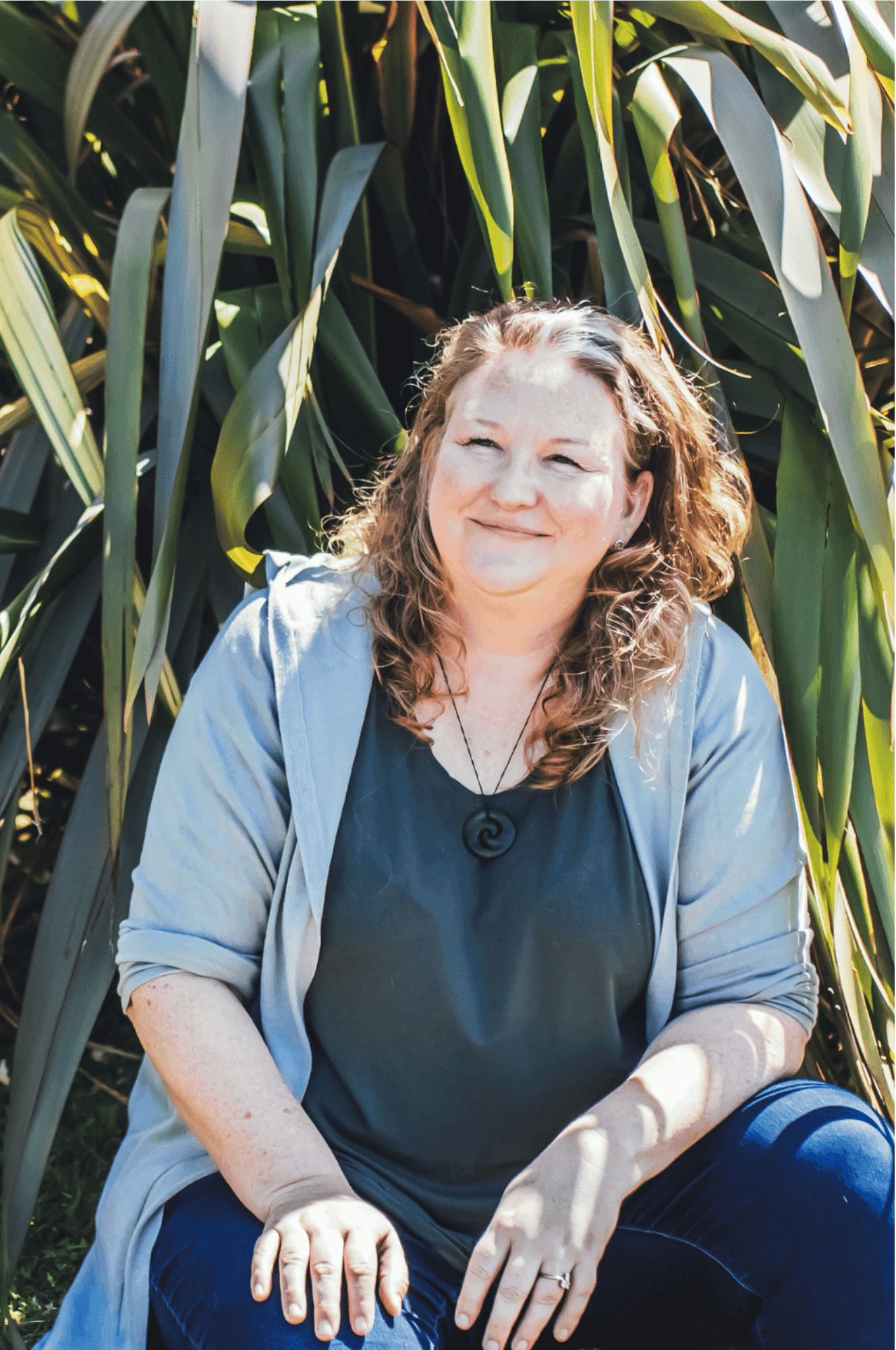 Photo of Elizabeth, ADHD Coaching expert, sitting on the ground looking away from the camera with a flax bush in the background.