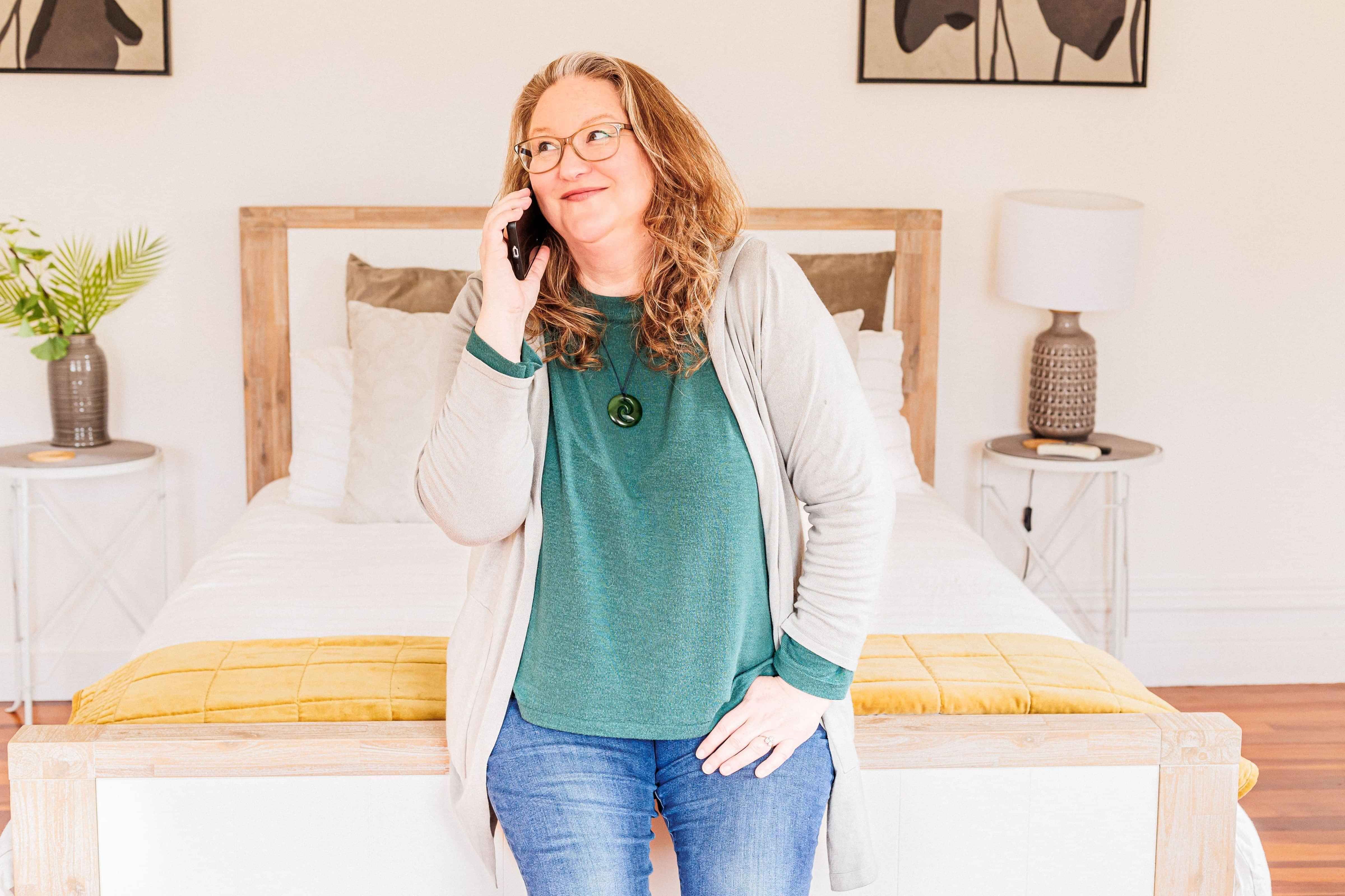 Photo of holistic health coaching expert, Elizabeth Marshall, sitting on the edge of a bed, looking away from the camera while on the phone.