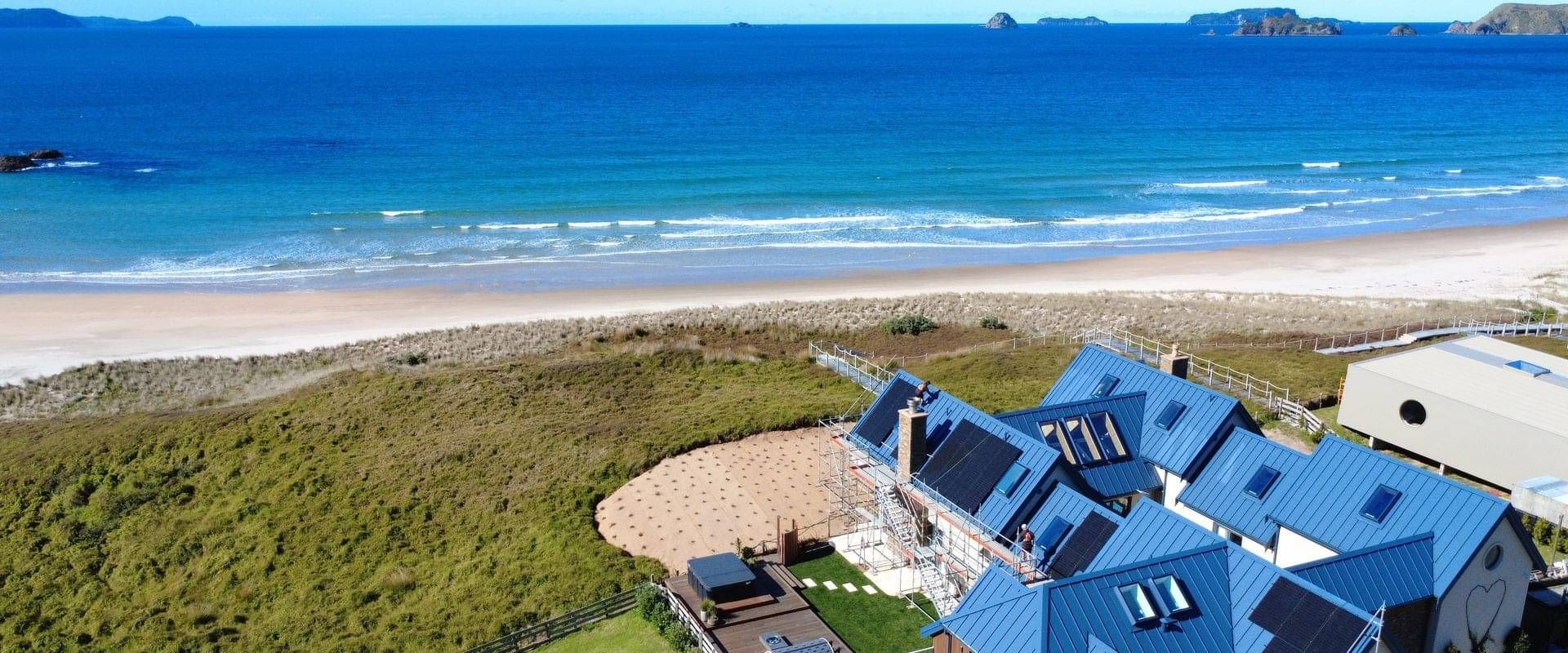 Aerial view of a blue-roofed beach house at Opito Bay with its new black solar panels, the beach and the offshore islands behind