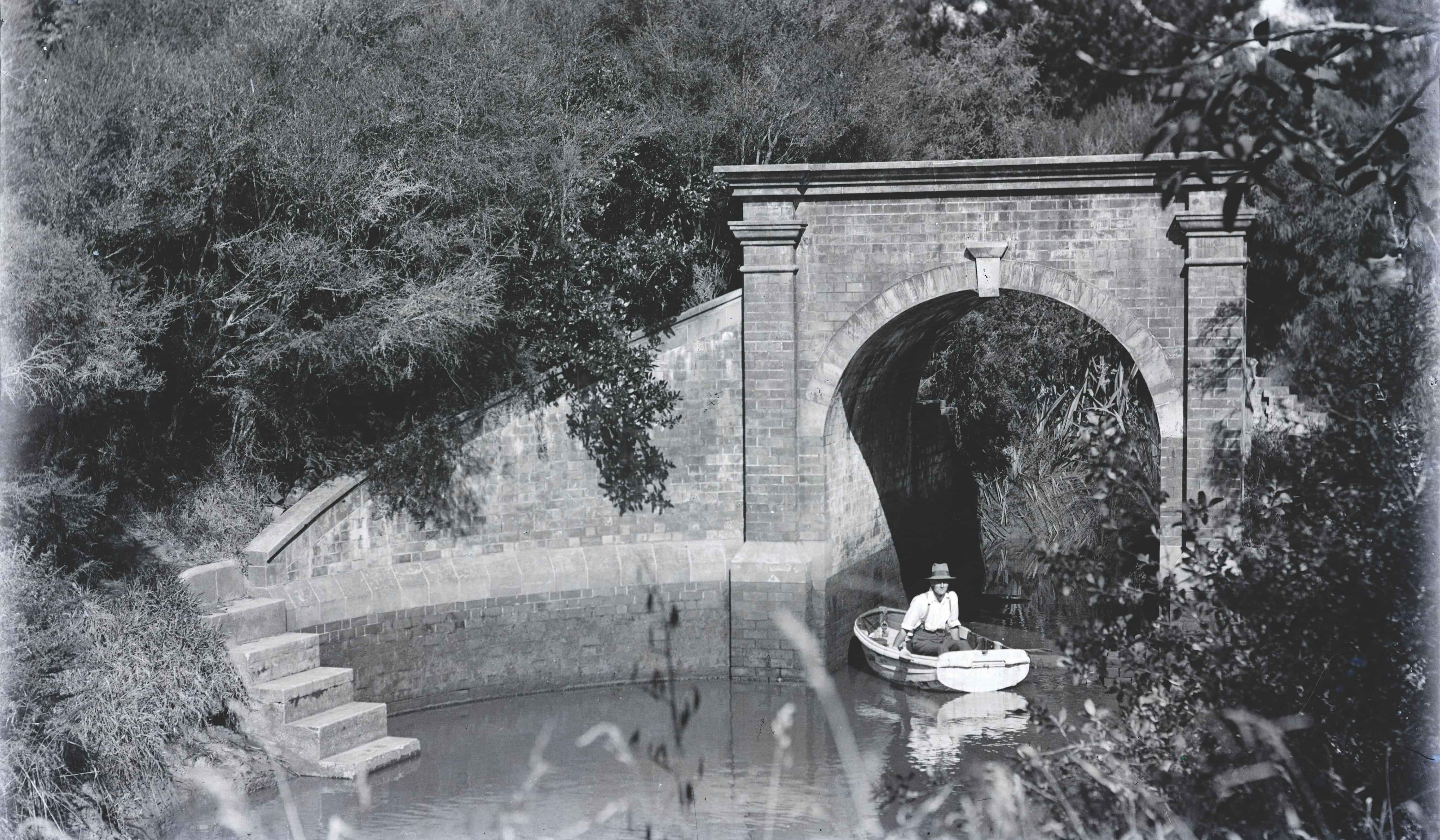 A man in a rowboat on a calm waterway with an arched bridge and bush in the background