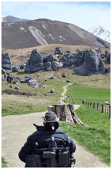 A man in an electric wheelchair on a path with a mountain range and large boulders ahead