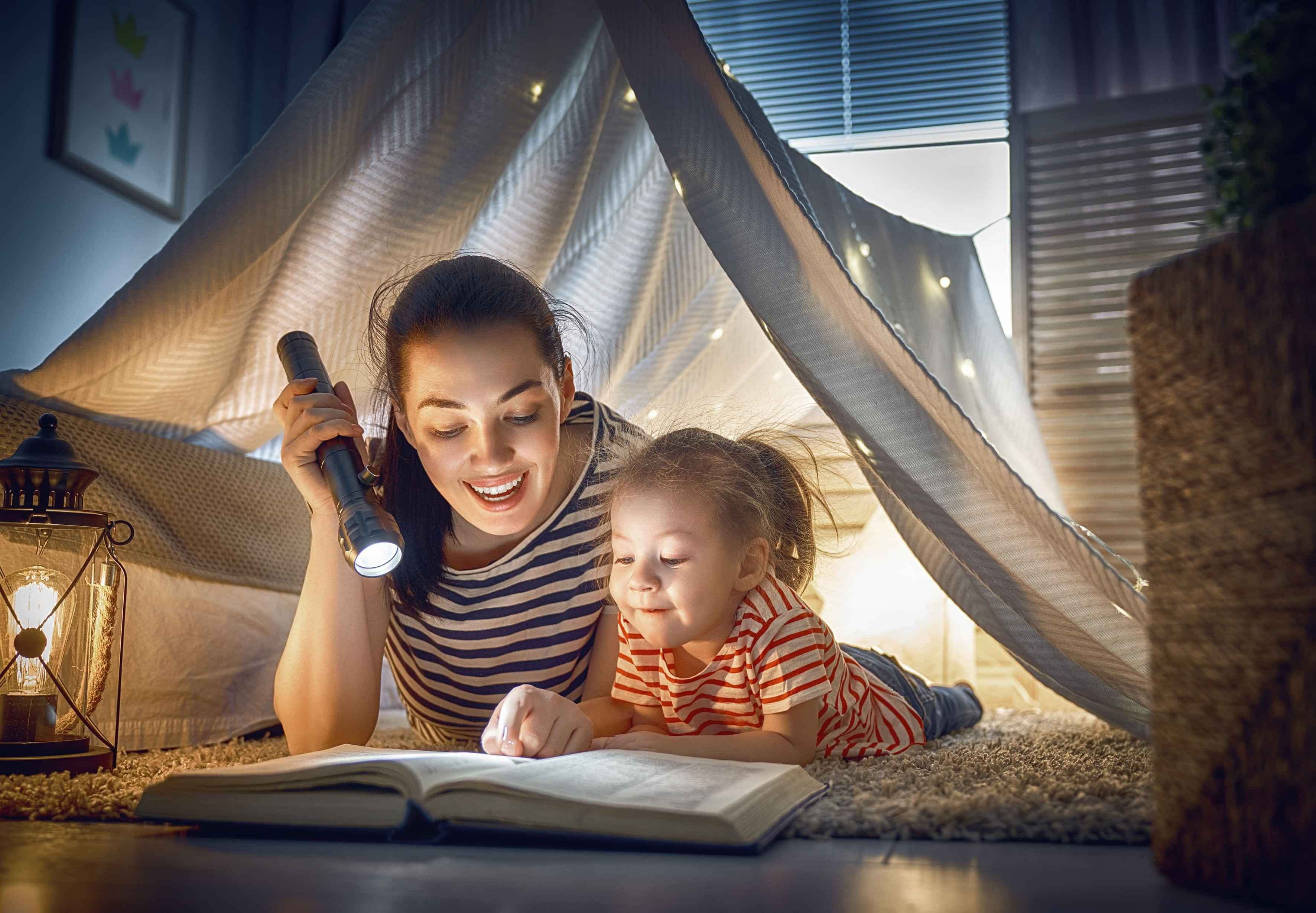 Mum and daughter in a sheet fort reading a book by torch