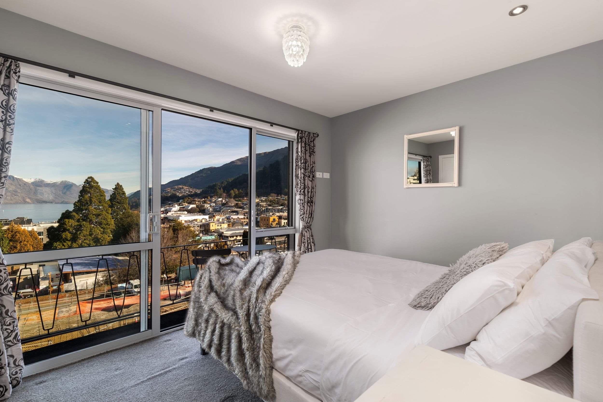 A beautifully staged master bedroom with white linen and a fur throw on the bed, looking out to views of Queenstown.