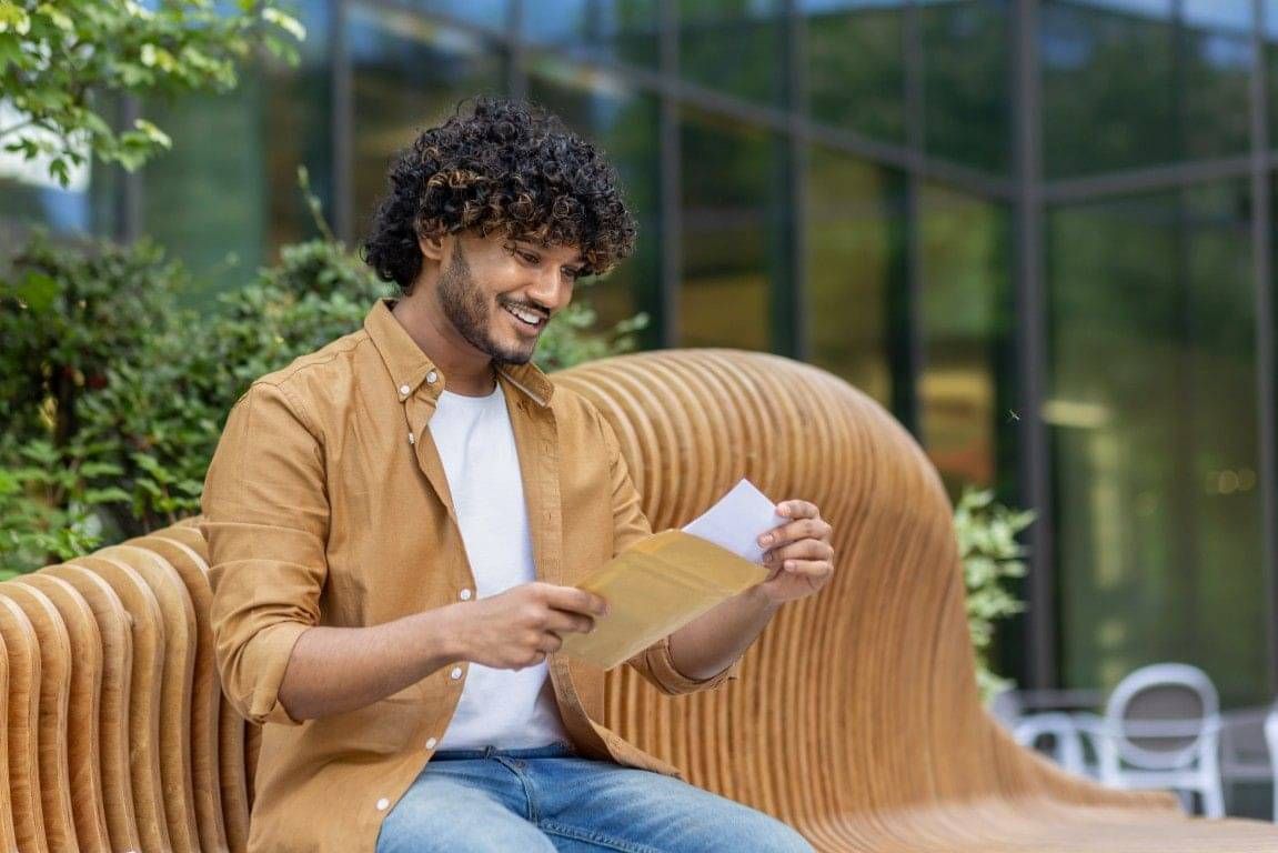 A smiling young Indian man is sitting on a bench outside and reading a report out of an envelope