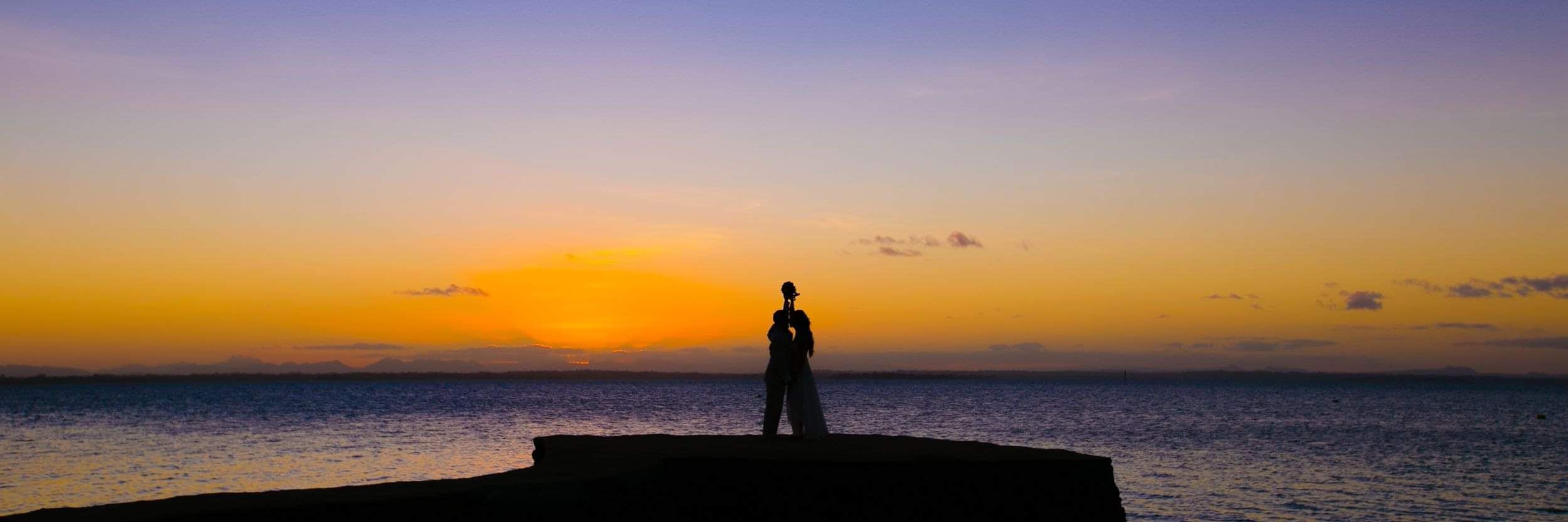Couple standing together at sunset overlooking the ocean, symbolising a destination wedding or romantic escape