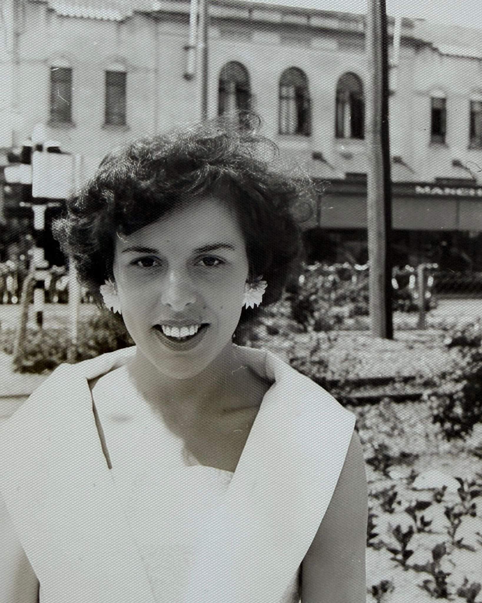 image of artist's mother smiling outdoors in a photograph from 1959, wearing a light sleeveless dress with buildings and gardens behind her