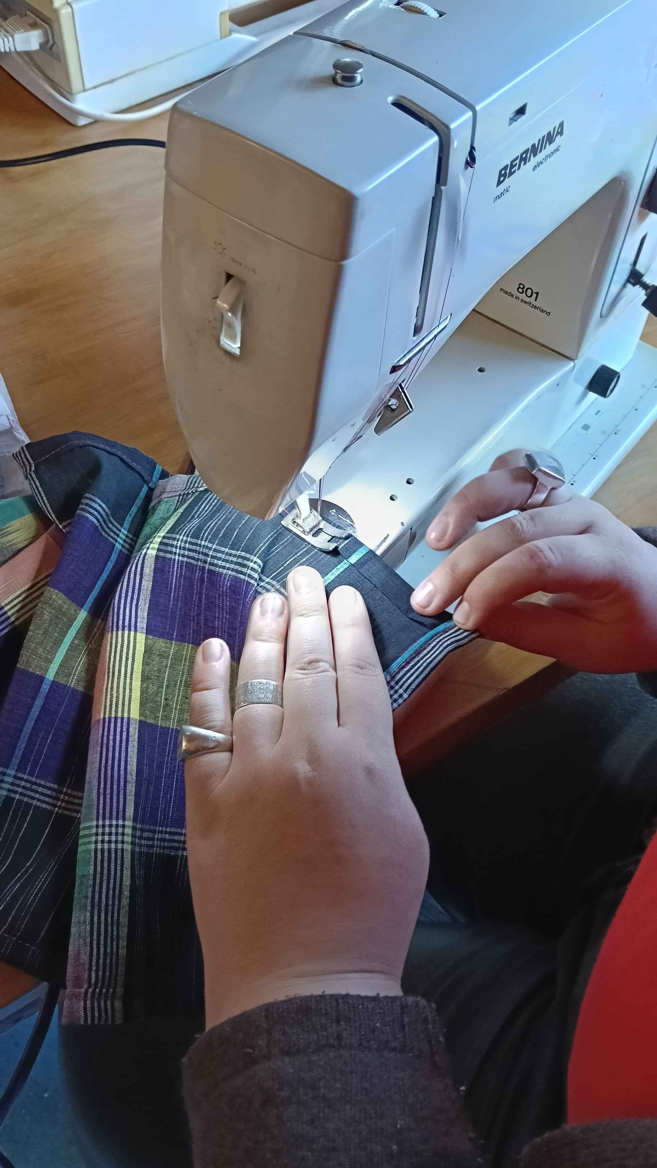 A close up shot of someone's hands feeding a blue tartan cloth through a sewing machine.