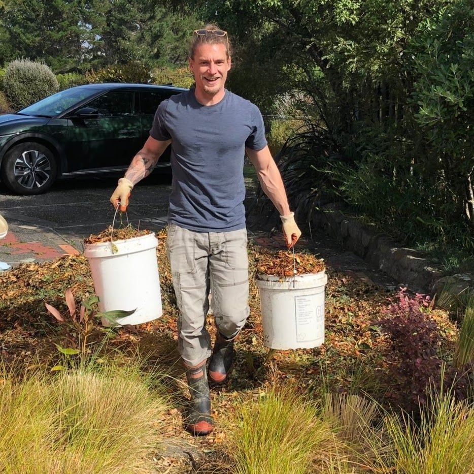 A man, shown from the chest down, carrying two buckets full of gardening mulch. He is wearing gumboots and walking through the grass.