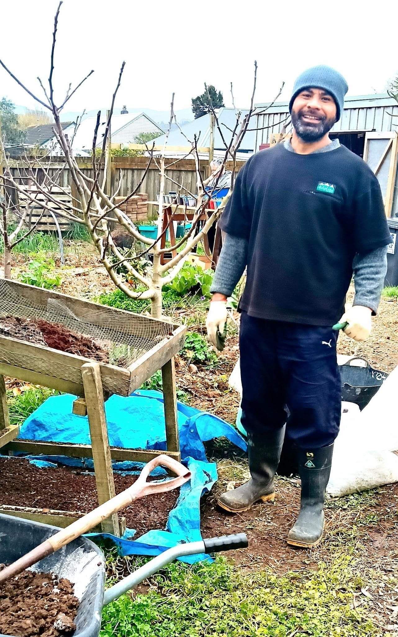 A timebanker smiling for a photo wearing gardening gloves next to a soil sifter.