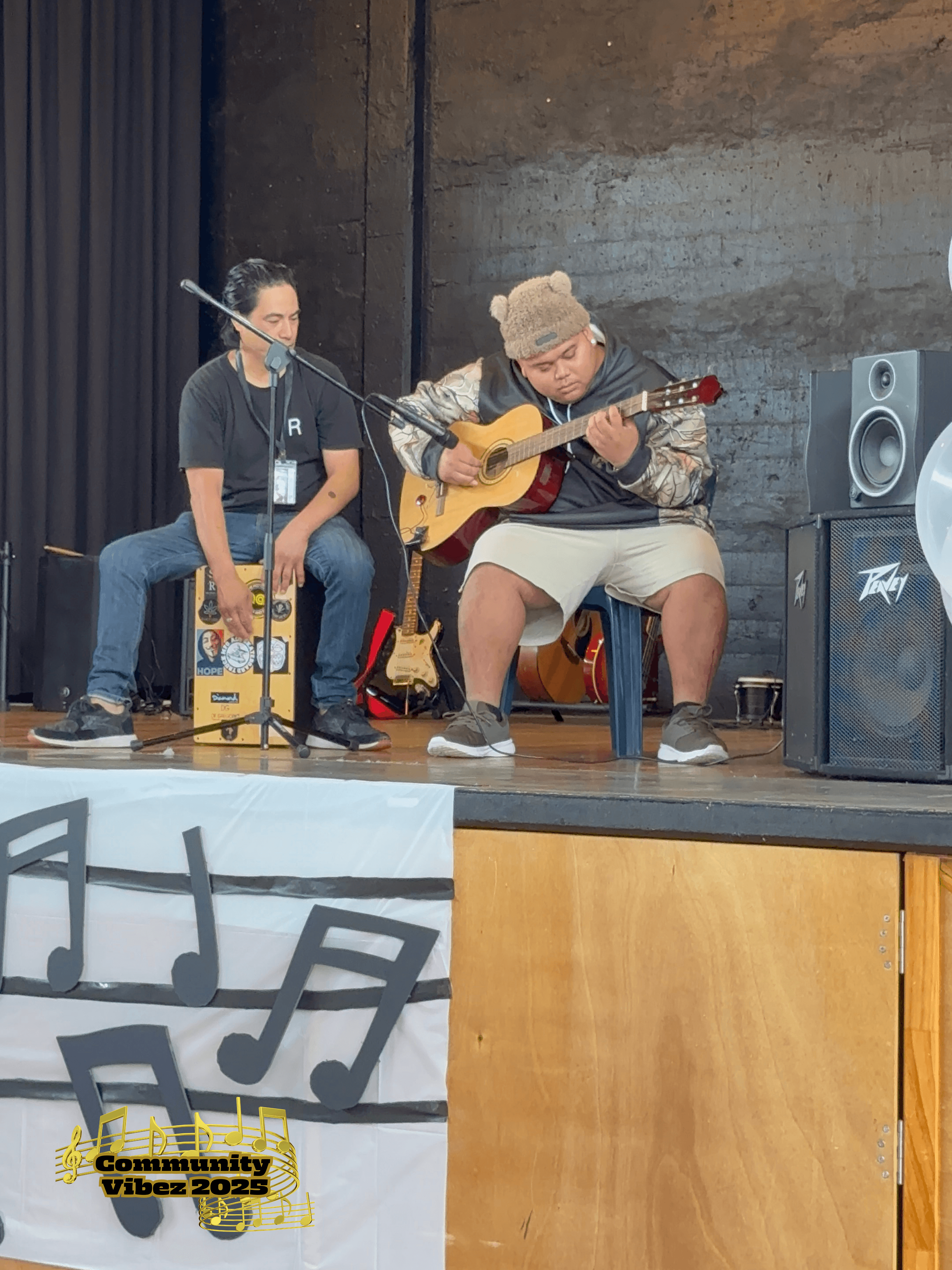 Two young men on stage. One is playing the guitar while the other watches them.