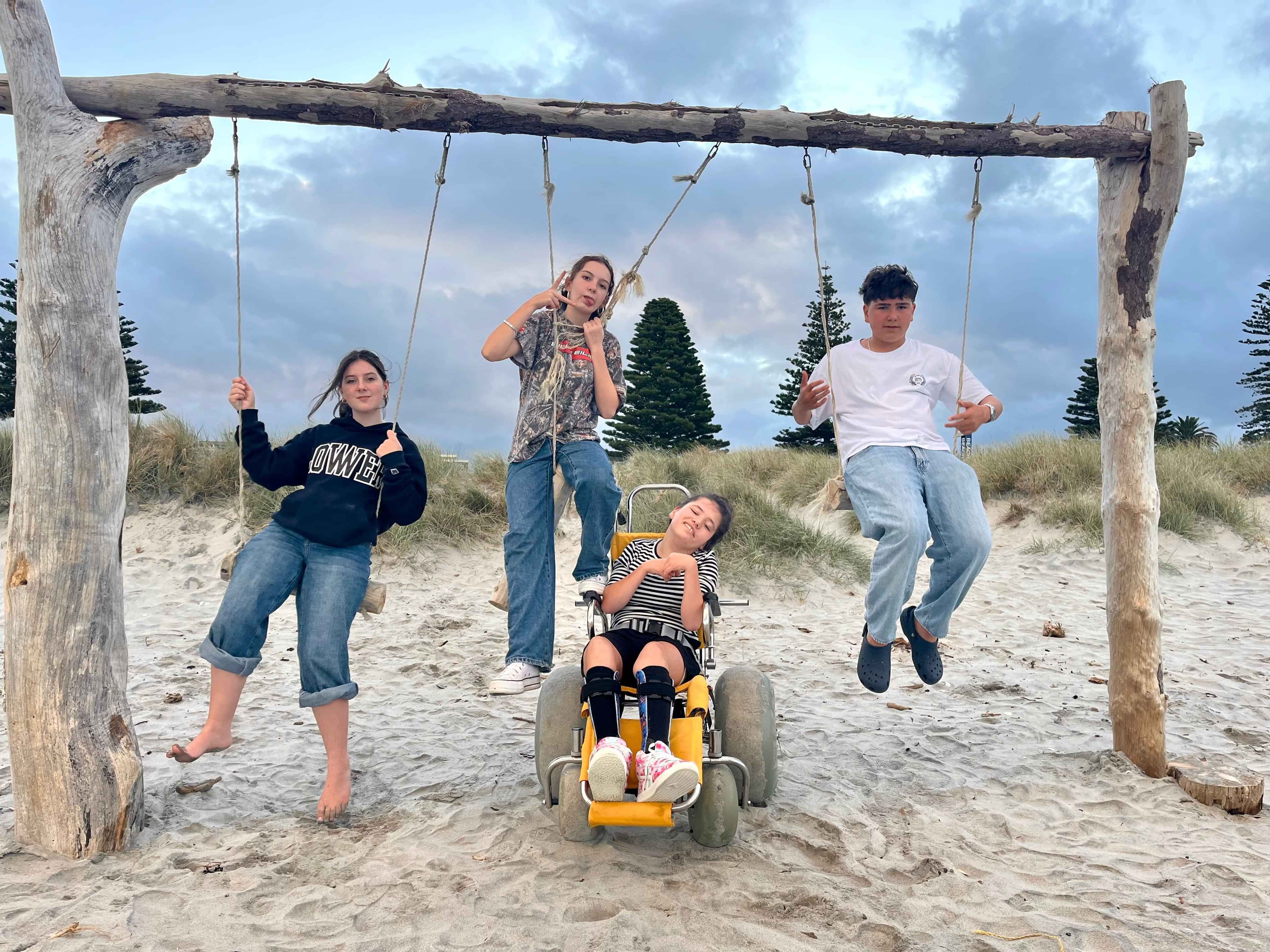 Four children are at the beach looking happy and carefree. Three sit on a driftwood swing and one is in front using a beach wheelchair. 