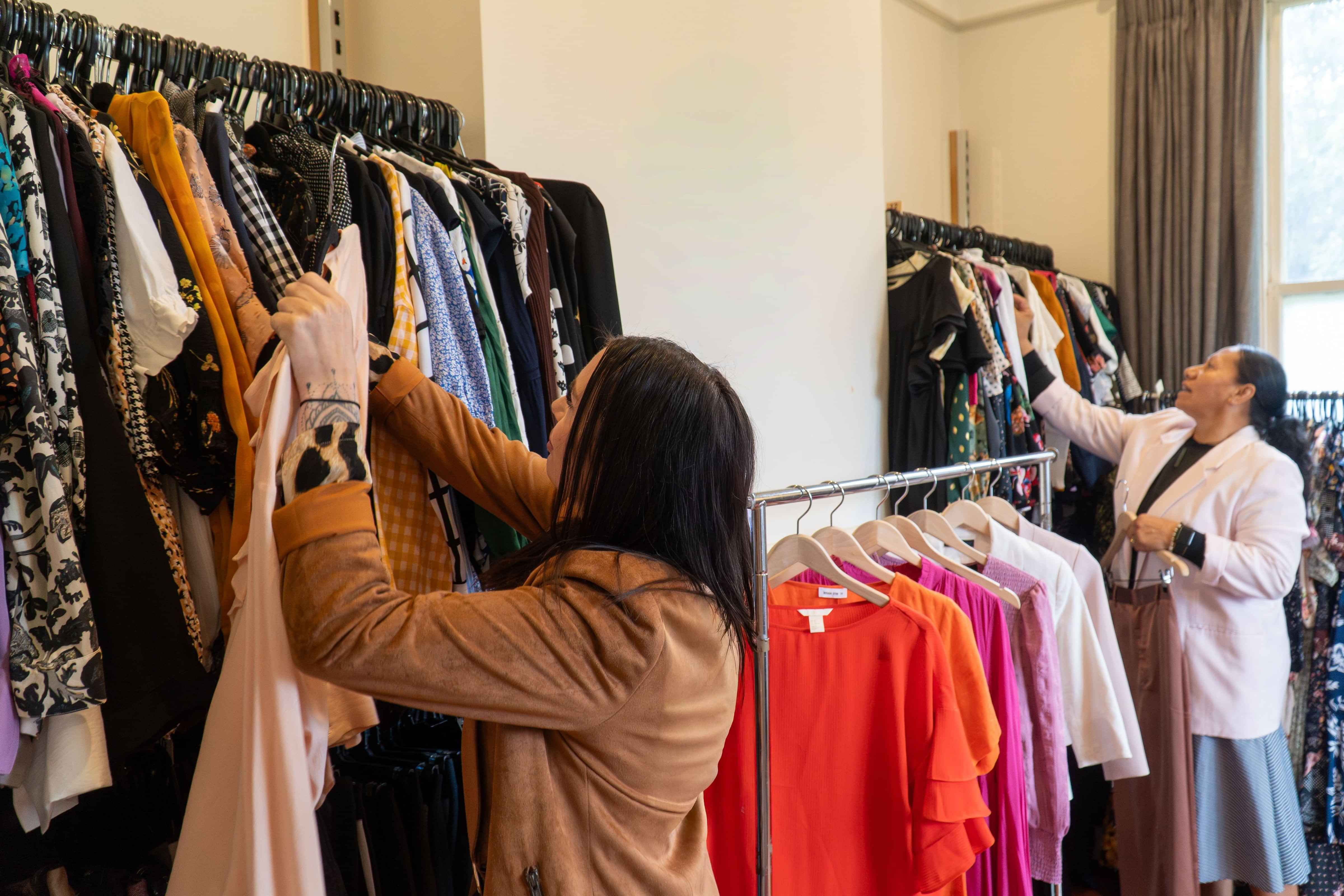 Two Dress for Success clients look through clothing racks around styling room and pull out clothes to try on that they like